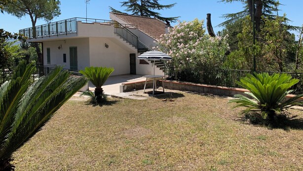 A two-story house with a balcony, a staircase, and a garden with palm trees and flowering plants. - Villa Panorama (Bivio Acquavella)