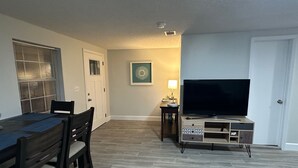 A living room with a TV, a dining table with chairs, a lamp, and a door. - Charming 2-bedroom cottage in lovely Fort Walton Beach (Fort Walton Beach)