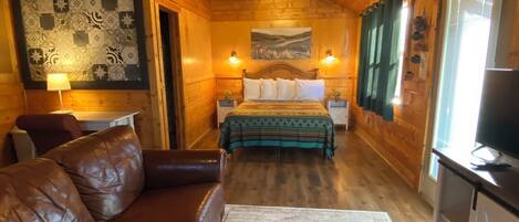A cozy cabin interior with a wooden ceiling, a bed with a green blanket, a leather sofa, a rug, and a TV.