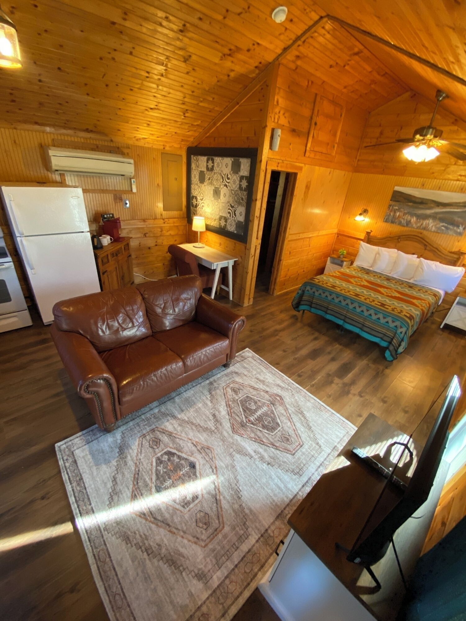 A cozy cabin interior with a wooden ceiling, a brown leather sofa, a small dining table, a bed with patterned bedding, and a television.
