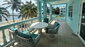 Een overdekt balkon met een witte tafel en rieten stoelen, uitkijkend op een strand met palmbomen en een pier.