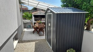 Un espacio exterior reducido con un cobertizo pequeño, una mesa con sillas y una planta en maceta.