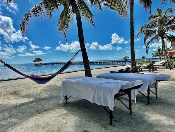 A beach with hammocks and sunbeds, a pier in the background, and palm trees.
