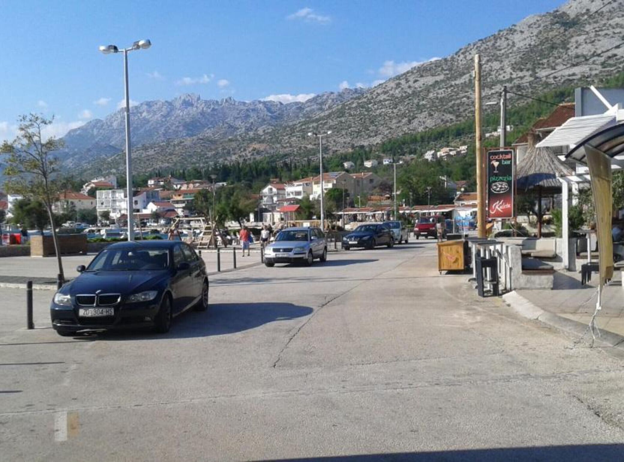 A street with cars parked on the side, a building with a signboard, and a mountainous backdrop.
