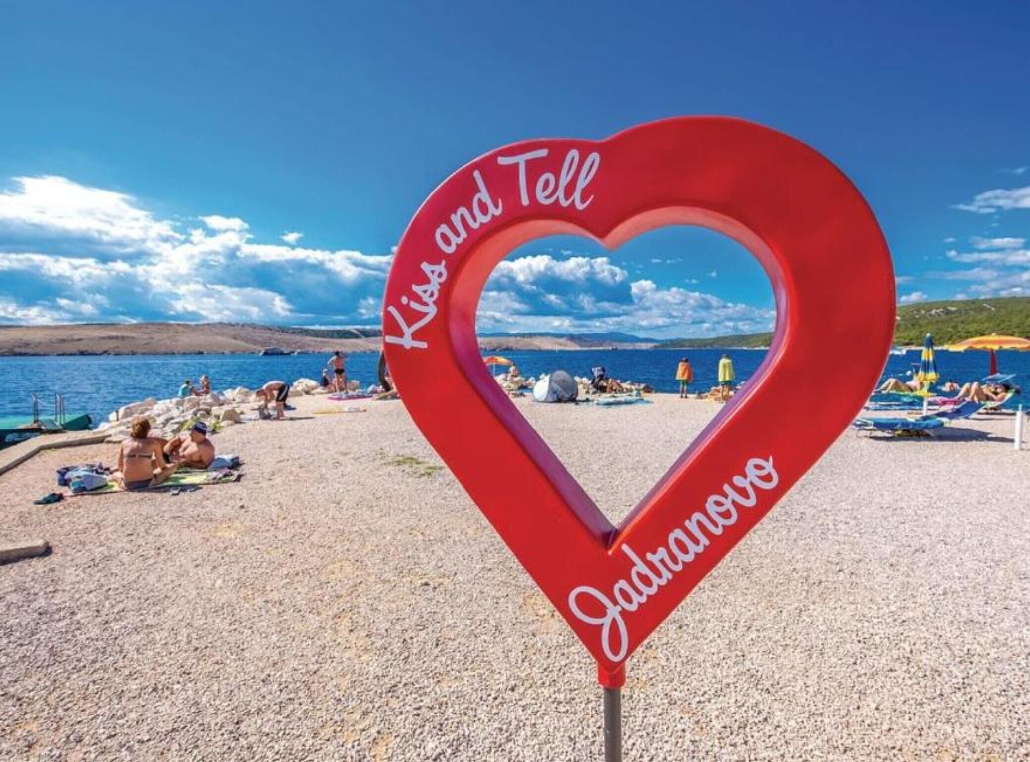 A heart-shaped sign with the words "Kiss and Tell" and "Jadransko" on a beach.