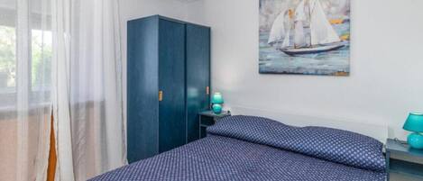 A bedroom with a bed, a wardrobe, a painting of sailboats, and a window with sheer curtains.