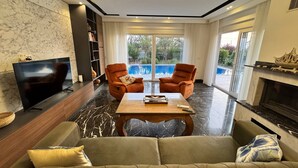 A modern living room with a sofa, two armchairs, a coffee table, a flat-screen TV, a fireplace, and a large window with a view of a swimming pool. - Entire villa in Keşefli, Turkey (Keşefli)