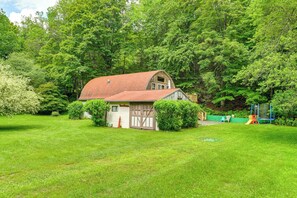 Une maison de style rustique, rappelant une grange, avec un toit rouge, entourée d’arbres verdoyants et d’une pelouse bien entretenue.