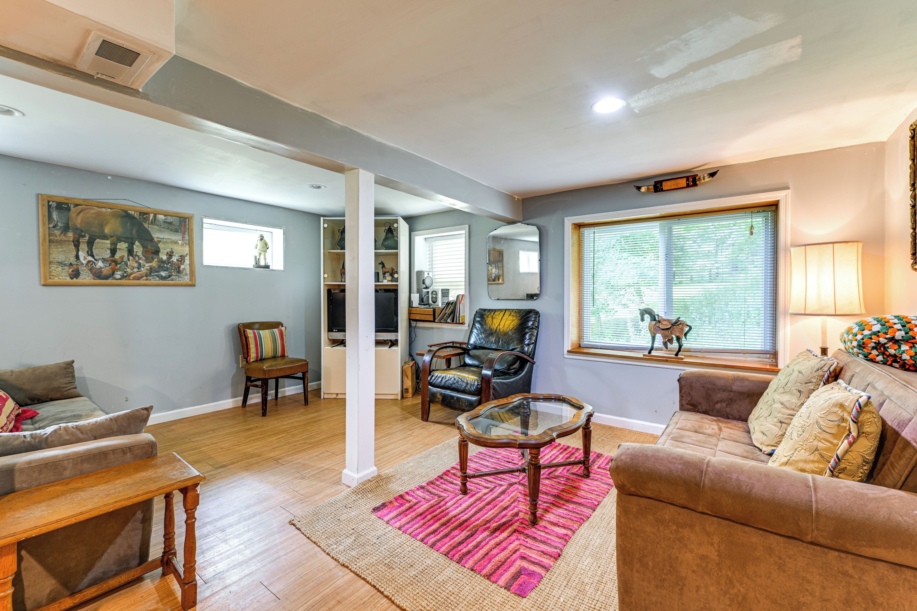A living room with a sofa, armchair, coffee table, and a rug. There is a window with blinds and a lamp on the right.