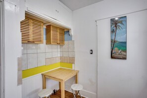 Ein kleiner Raum mit einem Holztisch, zwei Hocker, einem Fenster mit Holzjalousien und einem Strandbild an der Wand.
