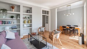 Un salon moderne avec un canapé, une table basse et des fauteuils en osier. On y trouve des étagères remplies de livres et d’objets décoratifs. Un coin repas avec une table et des chaises en bois est également visible.