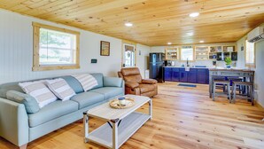 Een ruime woonkamer met een lichtblauwe bank, een bruine leren fauteuil en een houten salontafel. In de achtergrond is een keuken te zien met een blauw aanrecht en houten keukenkastjes.