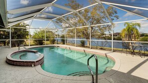 A covered outdoor swimming pool with a hot tub, surrounded by a patio area with tables and chairs, and a view of a lake and trees. - Pool & Dock! Canal-Front Port Charlotte Home (Port Charlotte)