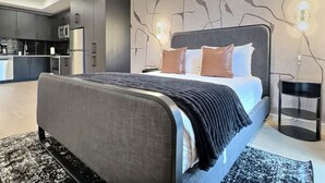 A modern bedroom with a large bed, a bedside table, and a nightstand. There is a kitchen area visible in the background. - Prime Bham Location, Walk to UAB, Park and Dining (Birmingham)