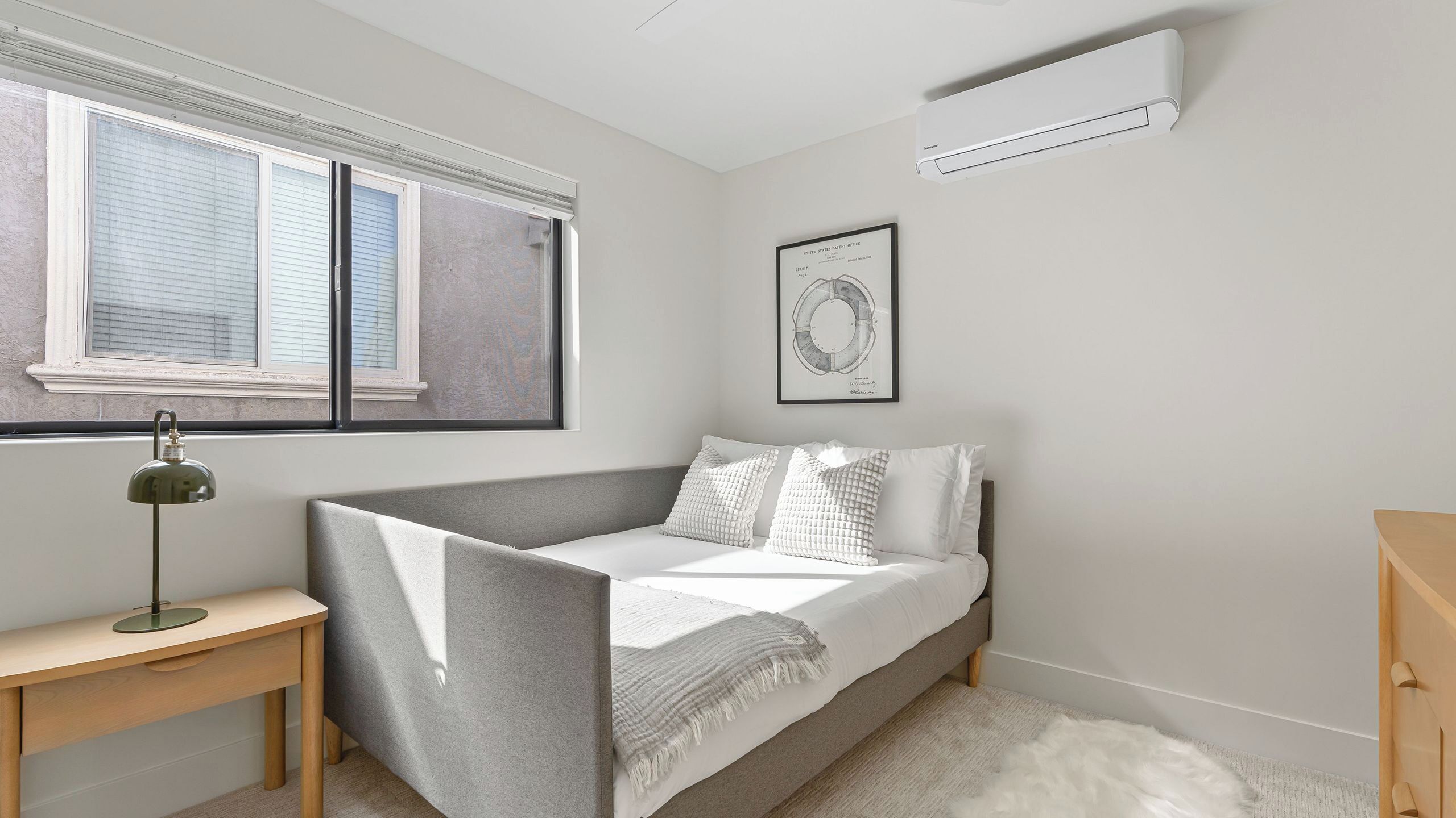 A bedroom with a bed, a desk, a lamp, a window, and an air conditioner.