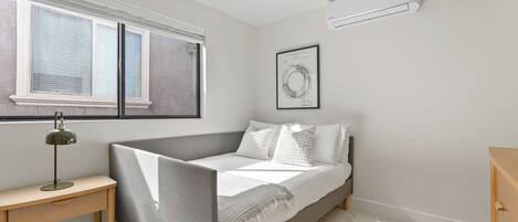 A bedroom with a bed, a desk, a lamp, a window, and an air conditioner.