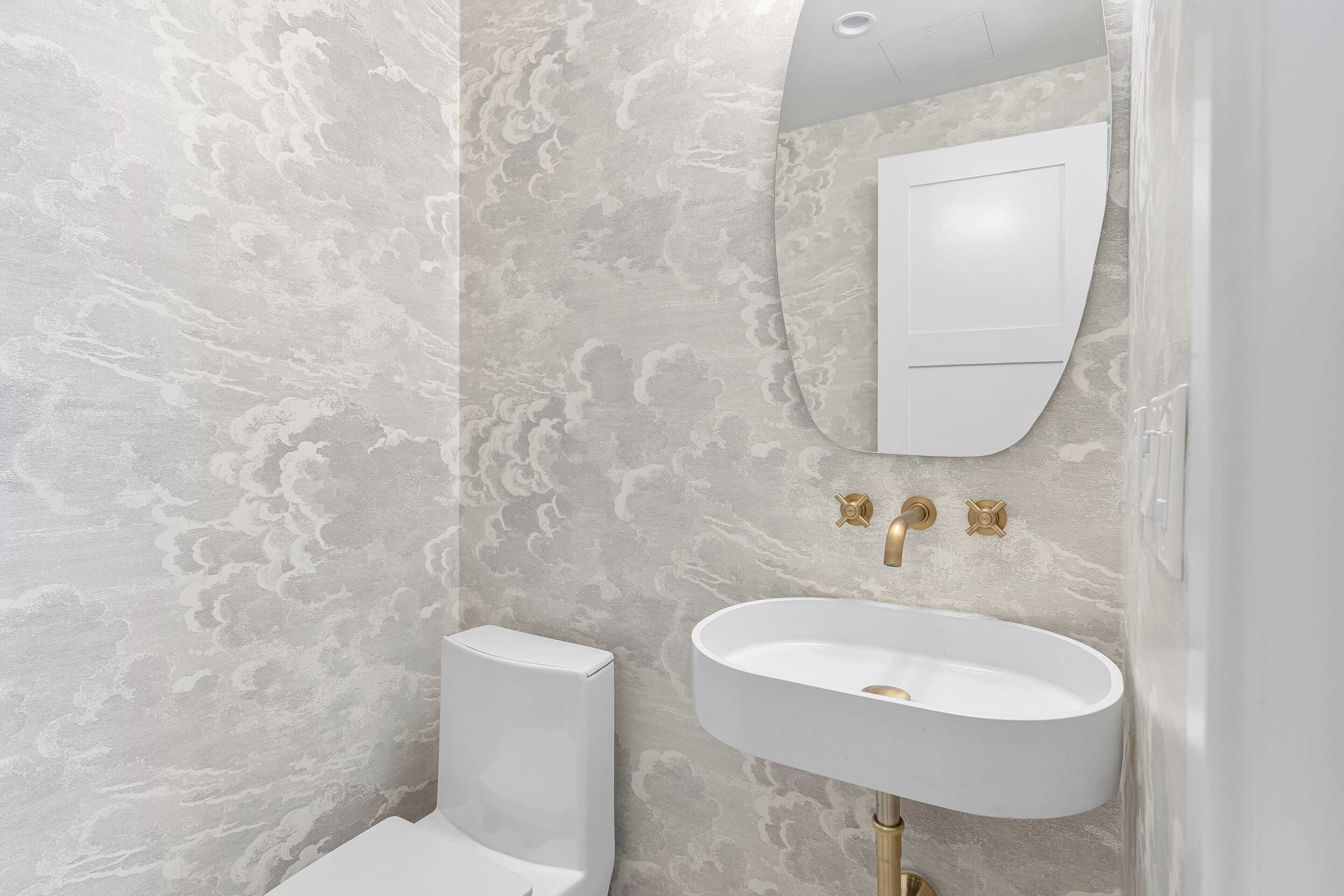 Een badkamer met een marmeren wand, een wit toilet, een witte wastafel met gouden details, en een spiegel boven de wastafel.