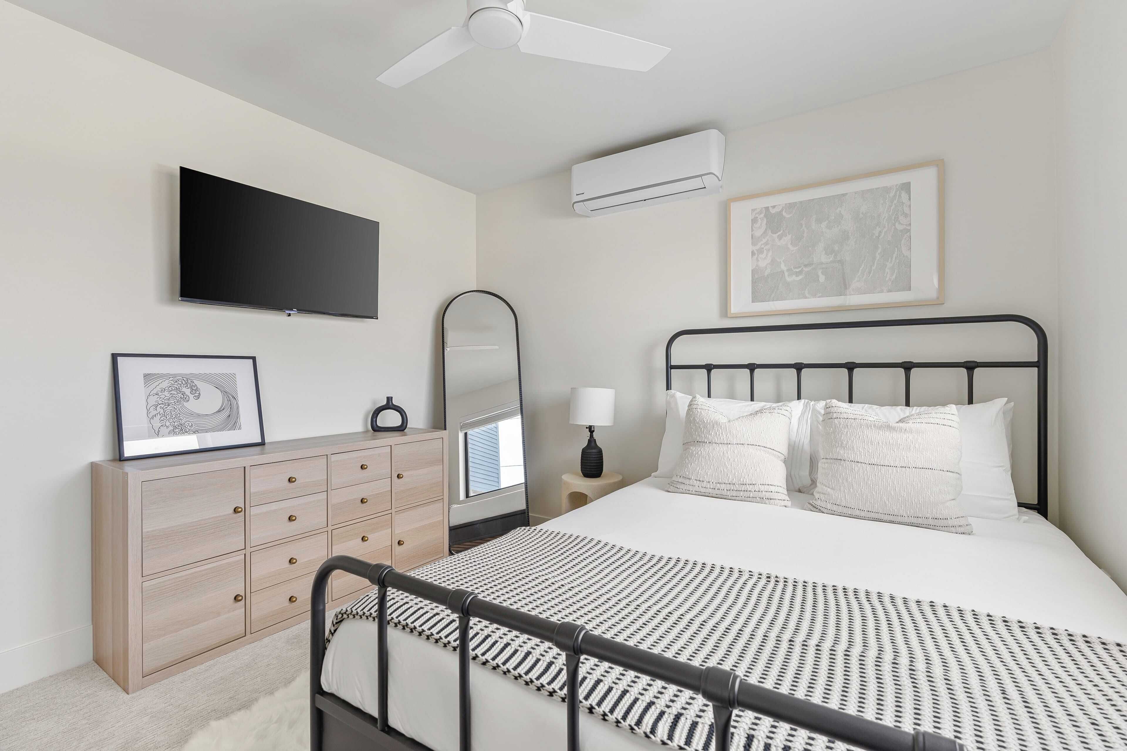 A bedroom with a bed, a dresser, a TV, a ceiling fan, an air conditioner, and a framed picture on the wall.
