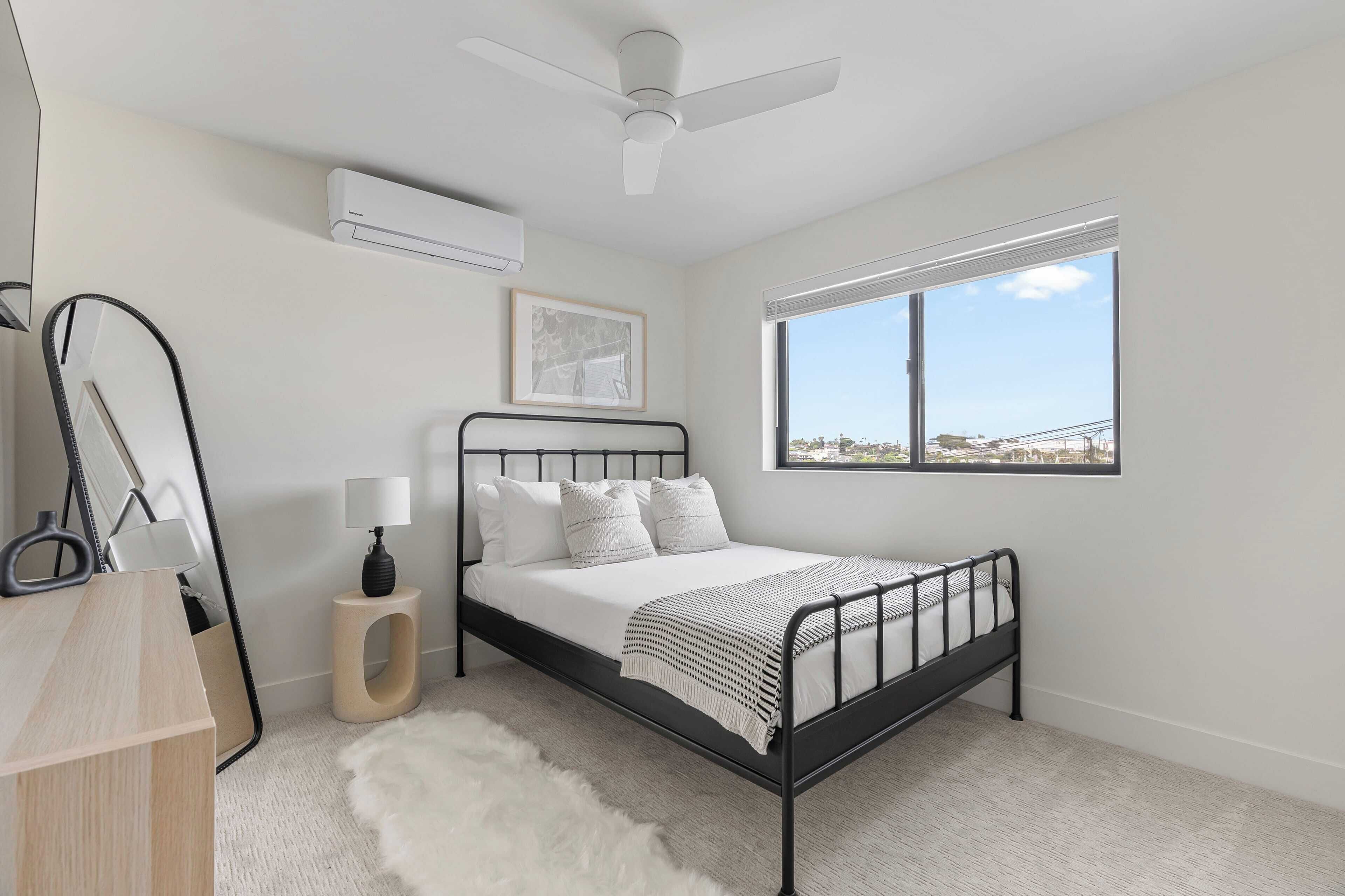 A bedroom with a bed, a ceiling fan, a window with a view, a small table with a lamp, and a white furry rug.