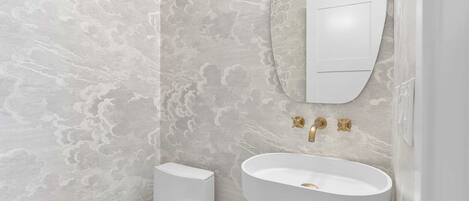 A bathroom with a marble wall, a white toilet, a white sink with gold fixtures, and a mirror above the sink.