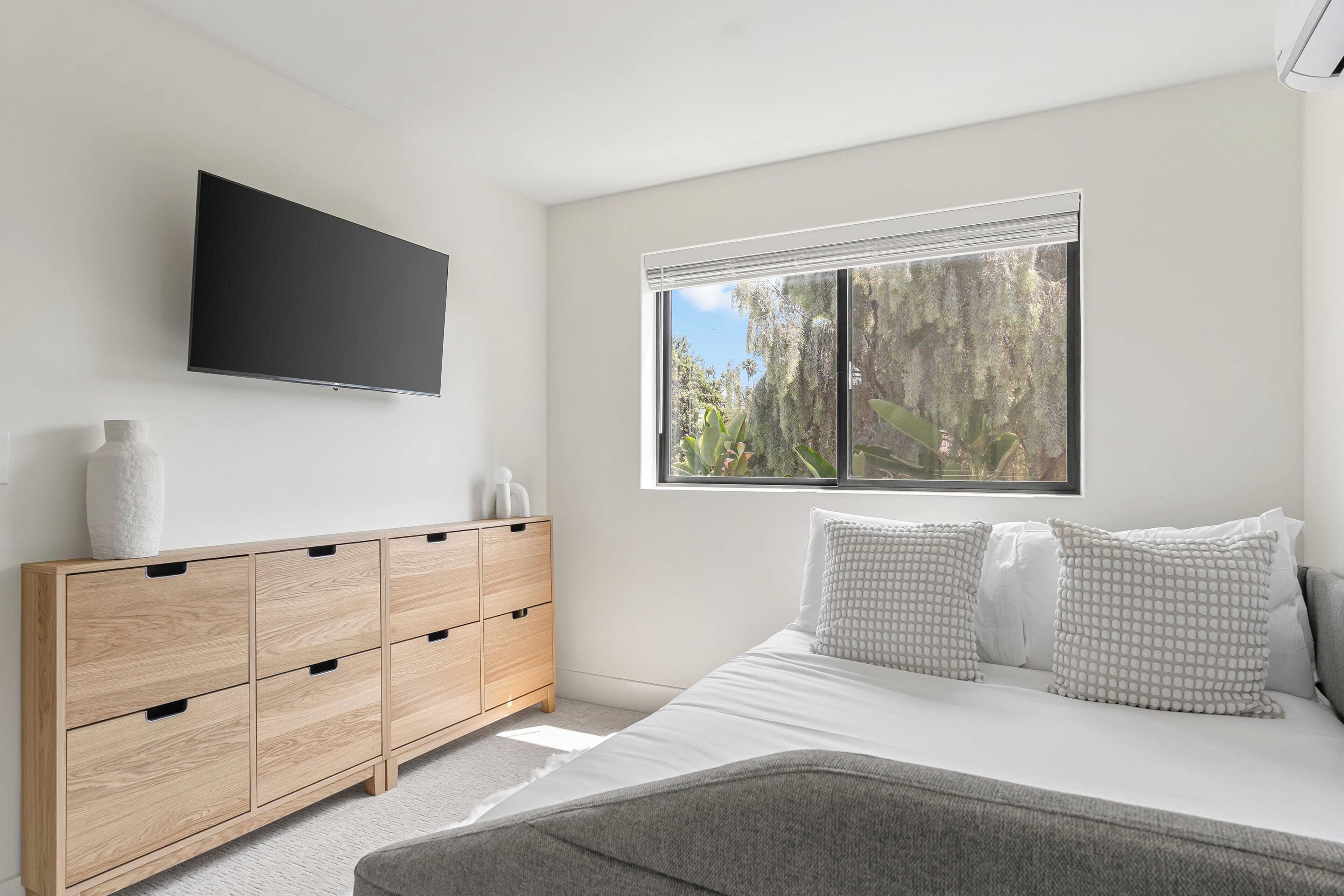 Una camera da letto con un letto, una cassettiera in legno, una TV a schermo piatto e una finestra vista alberi.