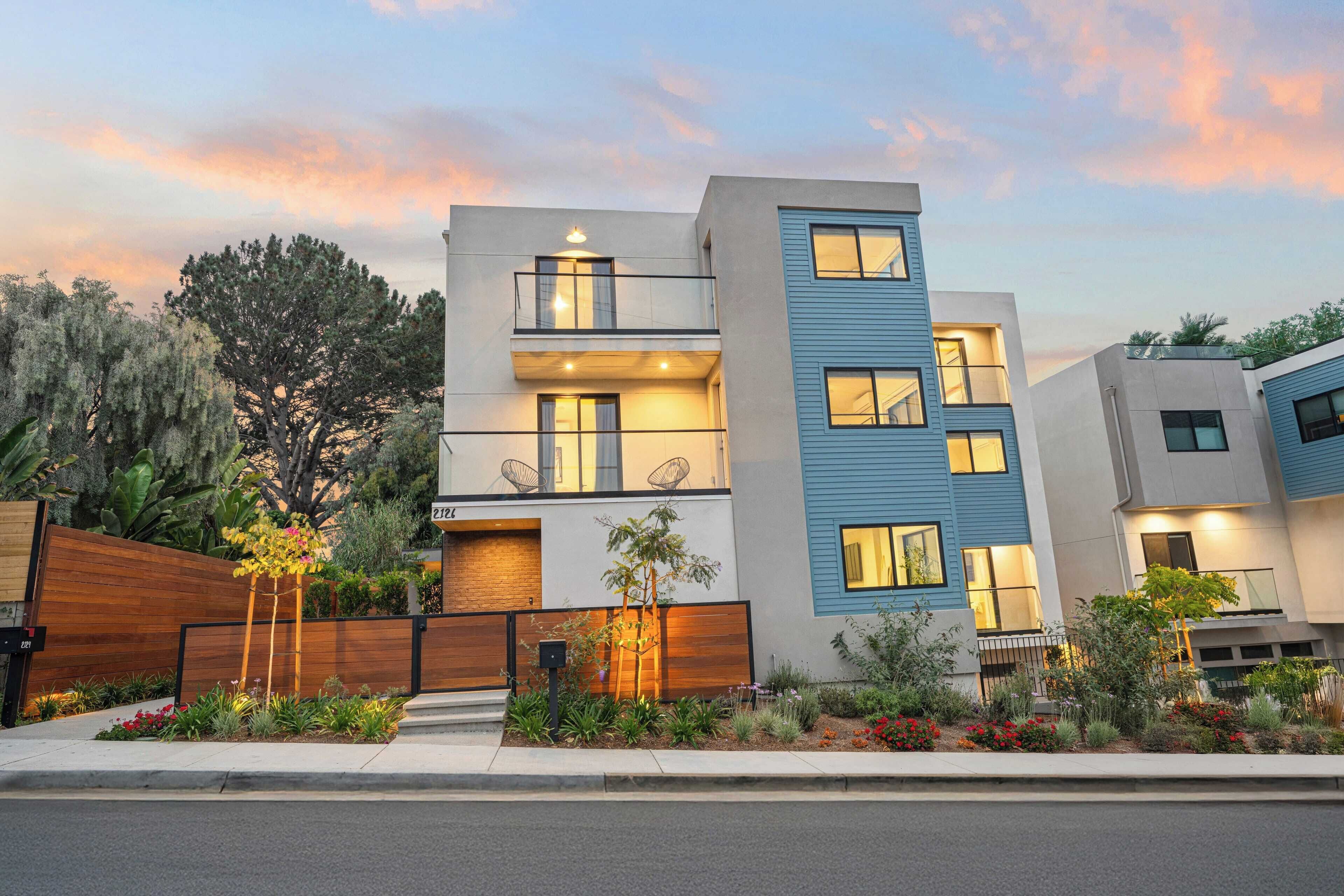 A modern multi-story residential building with a flat roof, large windows, and a well-maintained garden with various plants and flowers.