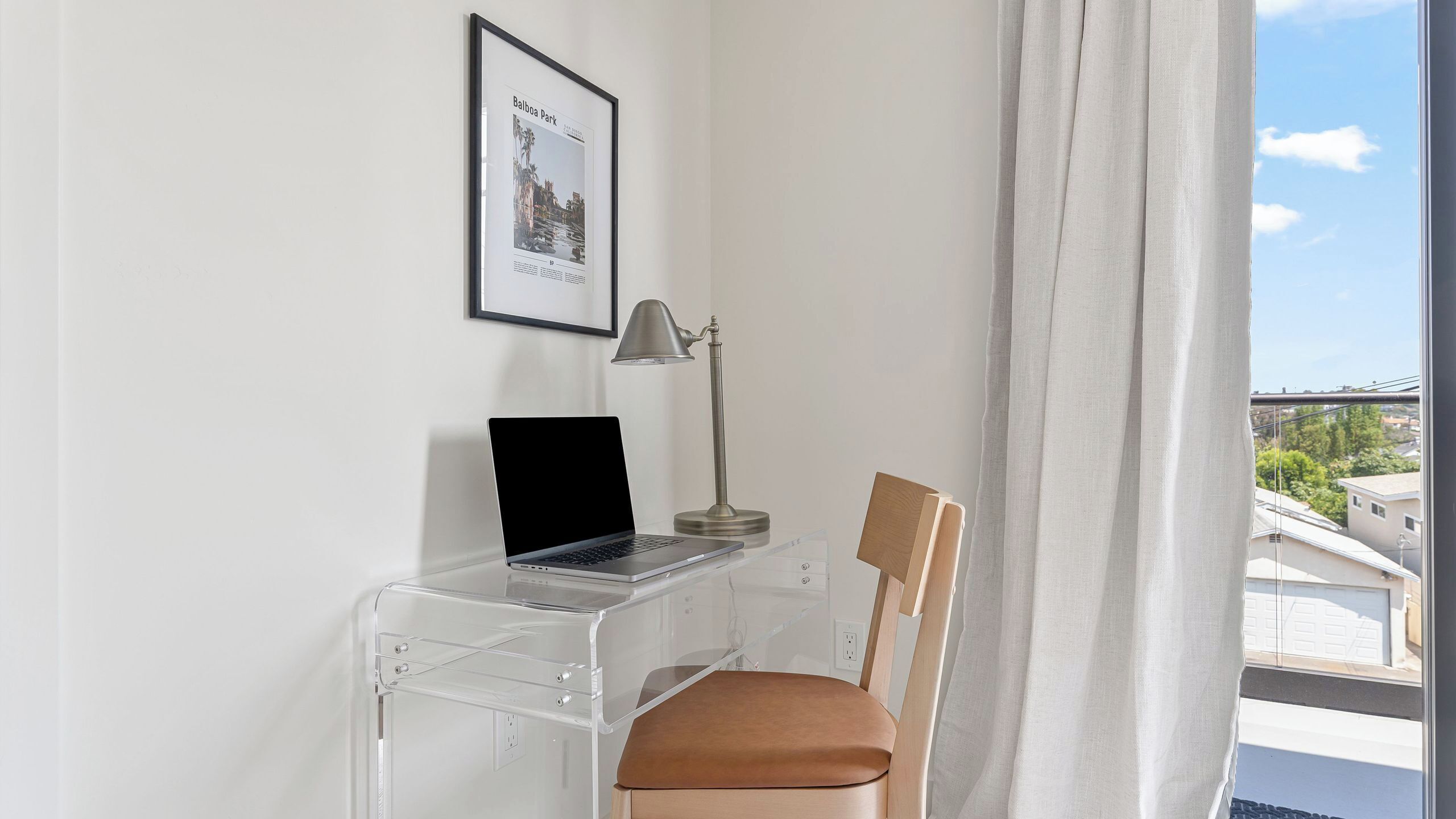 A clear desk with a laptop, a lamp, and a chair in a room with a window and a framed picture on the wall.