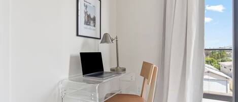 A clear desk with a laptop, a lamp, and a chair in a room with a window and a framed picture on the wall.