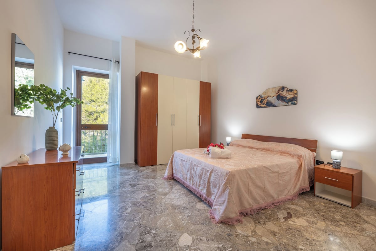 A bedroom with a bed, bedside table, nightstand, wardrobe, and a large window.