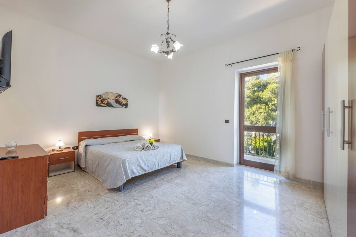 A bedroom with a bed, bedside table, lamp, and a view of the outdoors through a sliding door.