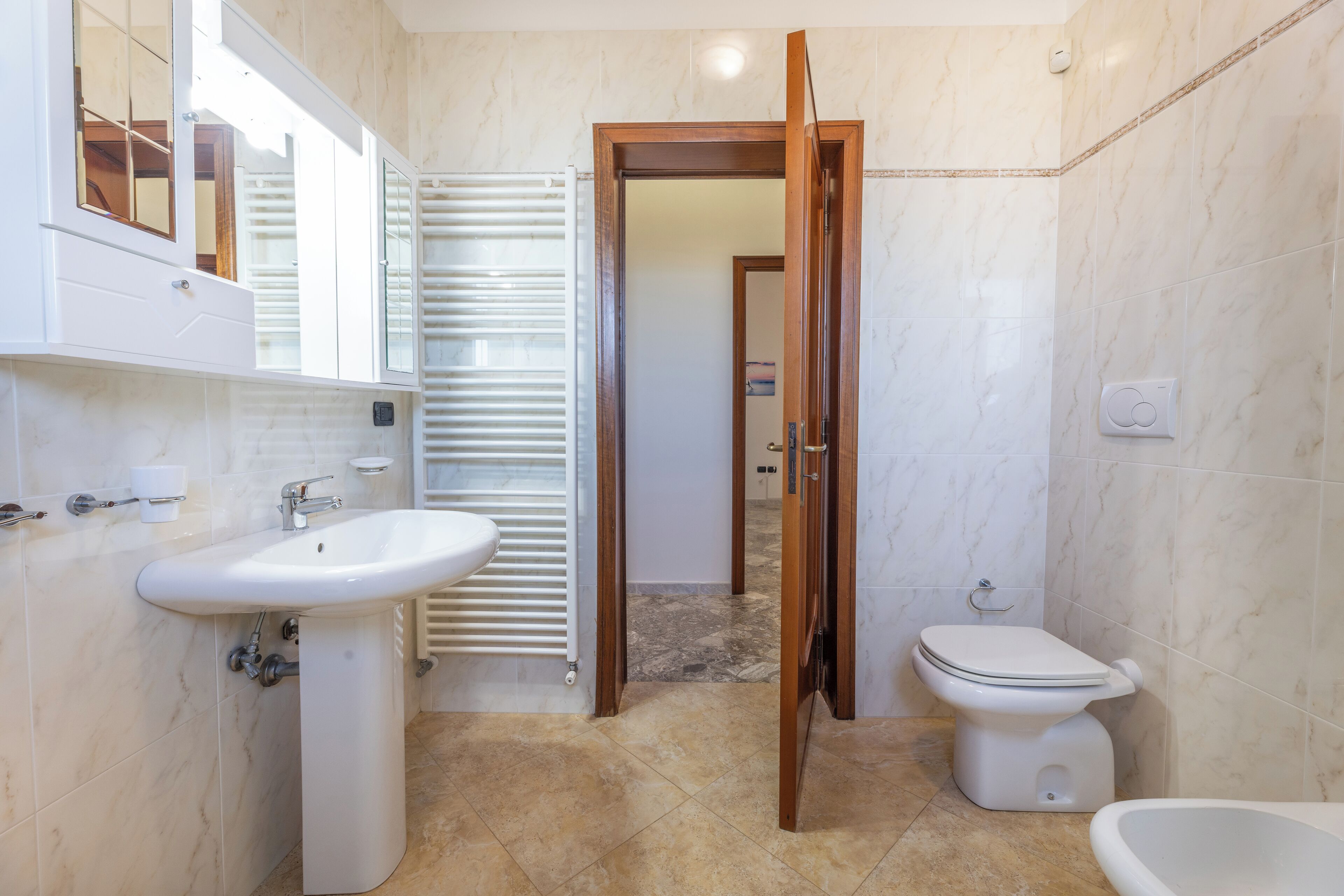 A bathroom with a white sink, toilet, and bidet, marble flooring, and a wooden door.