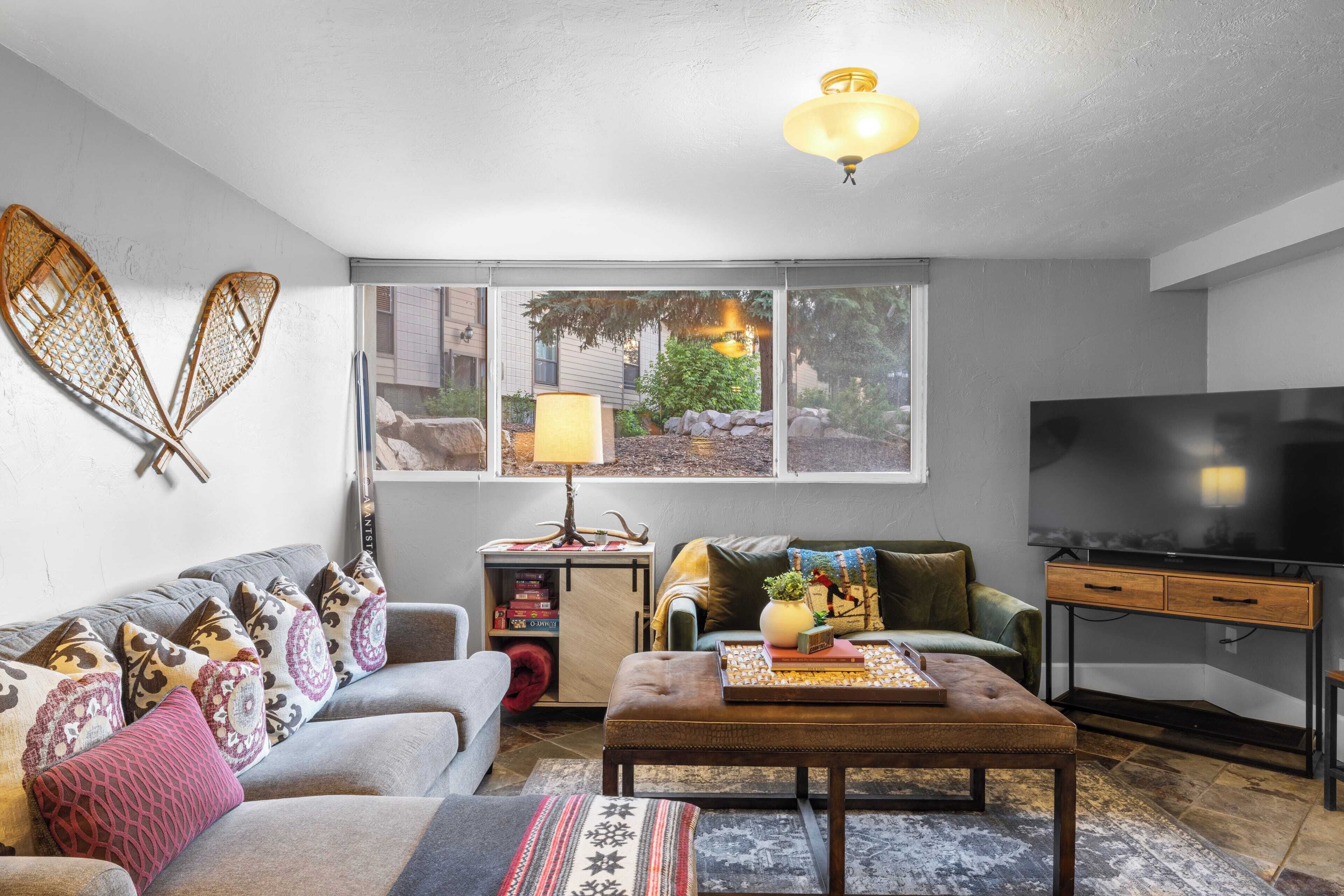 A living room with a sofa, ottoman, and coffee table, a television, and a window with a view of a garden.