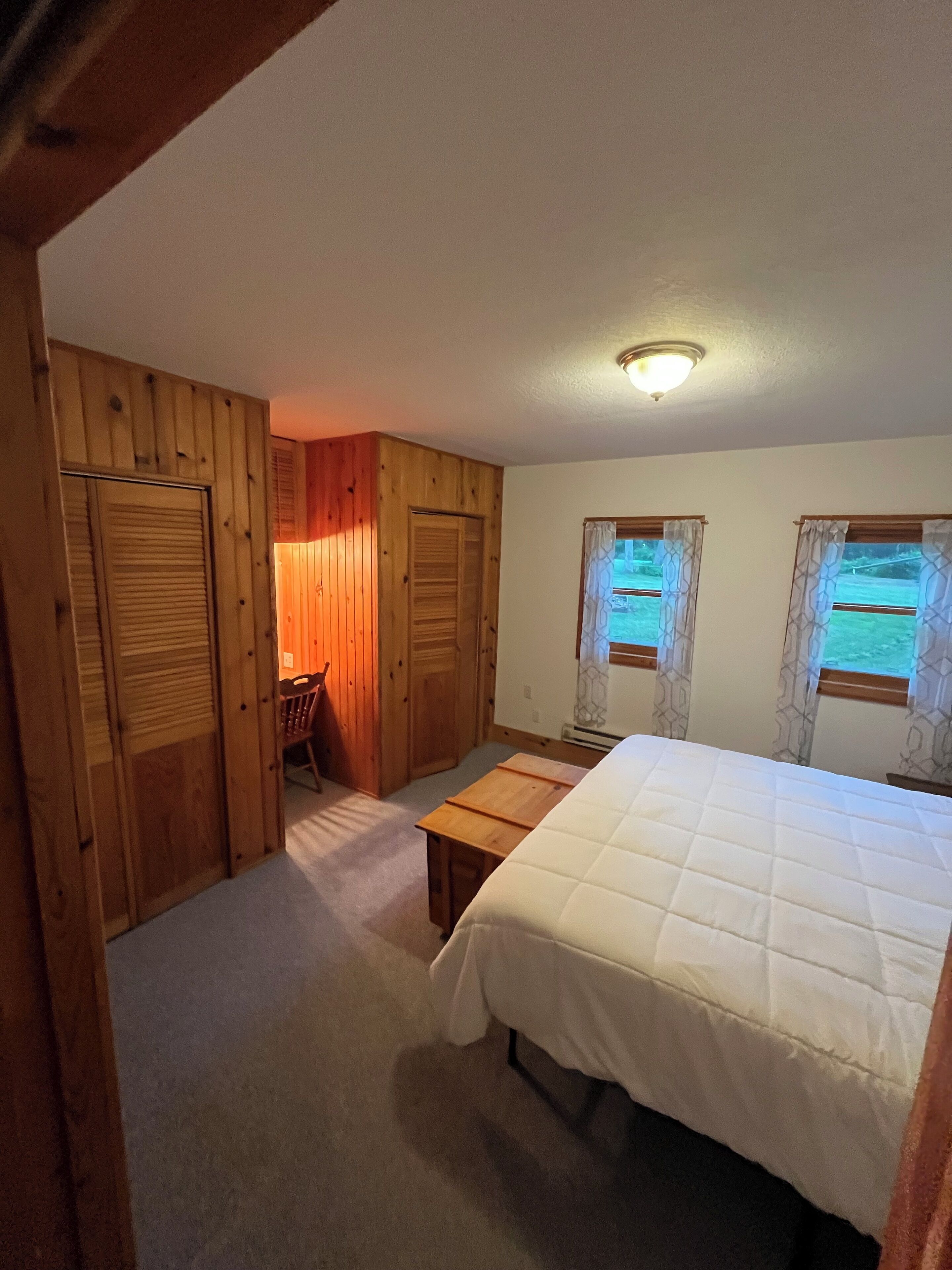 A bedroom with wooden walls, a bed, a small table, and two windows with curtains.