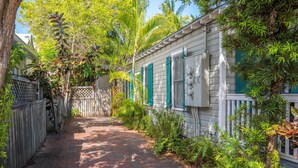 A narrow brick pathway flanked by lush greenery and a white building with green shutters.