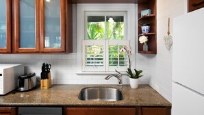 A kitchen with a stainless steel sink, a toaster, and a knife block. There is a window with blinds, a potted plant, and a shelf with decorative items.