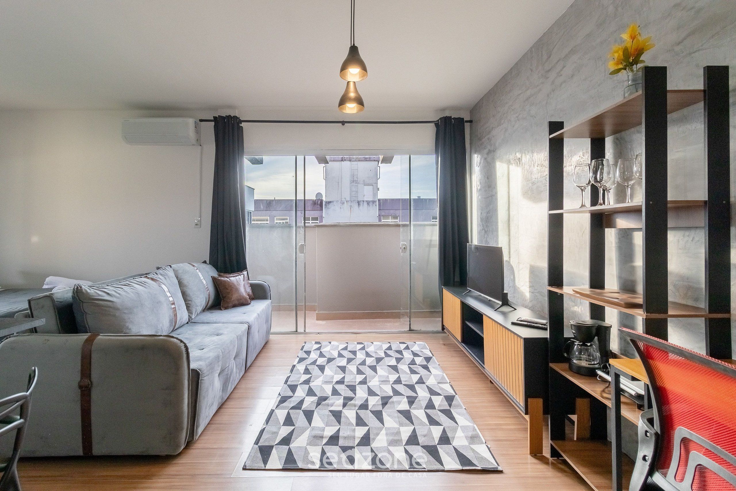 A modern living room with a grey sofa, a patterned rug, and a TV stand with a red chair.