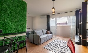 A modern living room with a green wall, a grey sofa, a patterned rug, and a dining area with chairs. - Fully Furnished Apartments 650m From Coqueiros Park - DAC (Florianópolis)