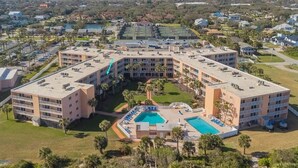 A multi-story hotel with a central courtyard featuring a swimming pool, surrounded by palm trees and a tennis court.