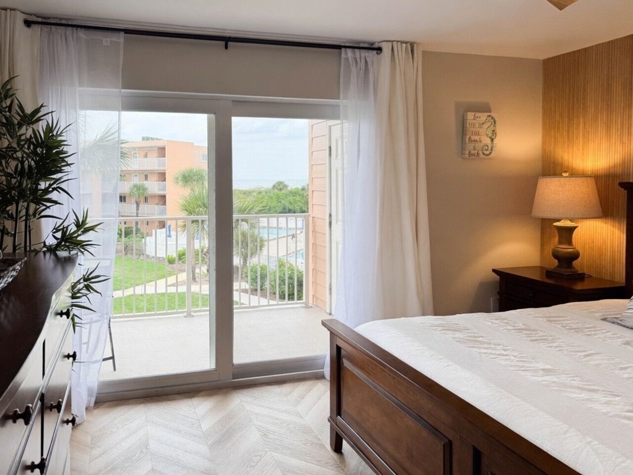 A hotel room with a bed, a nightstand, a lamp, a sliding glass door to a balcony, and a potted plant.