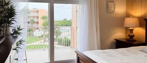 A hotel room with a bed, a nightstand, a lamp, a sliding glass door to a balcony, and a potted plant.