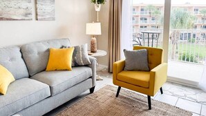 A living room with a grey sofa, a yellow armchair, a small side table with a lamp, and a view of a balcony with outdoor furniture.