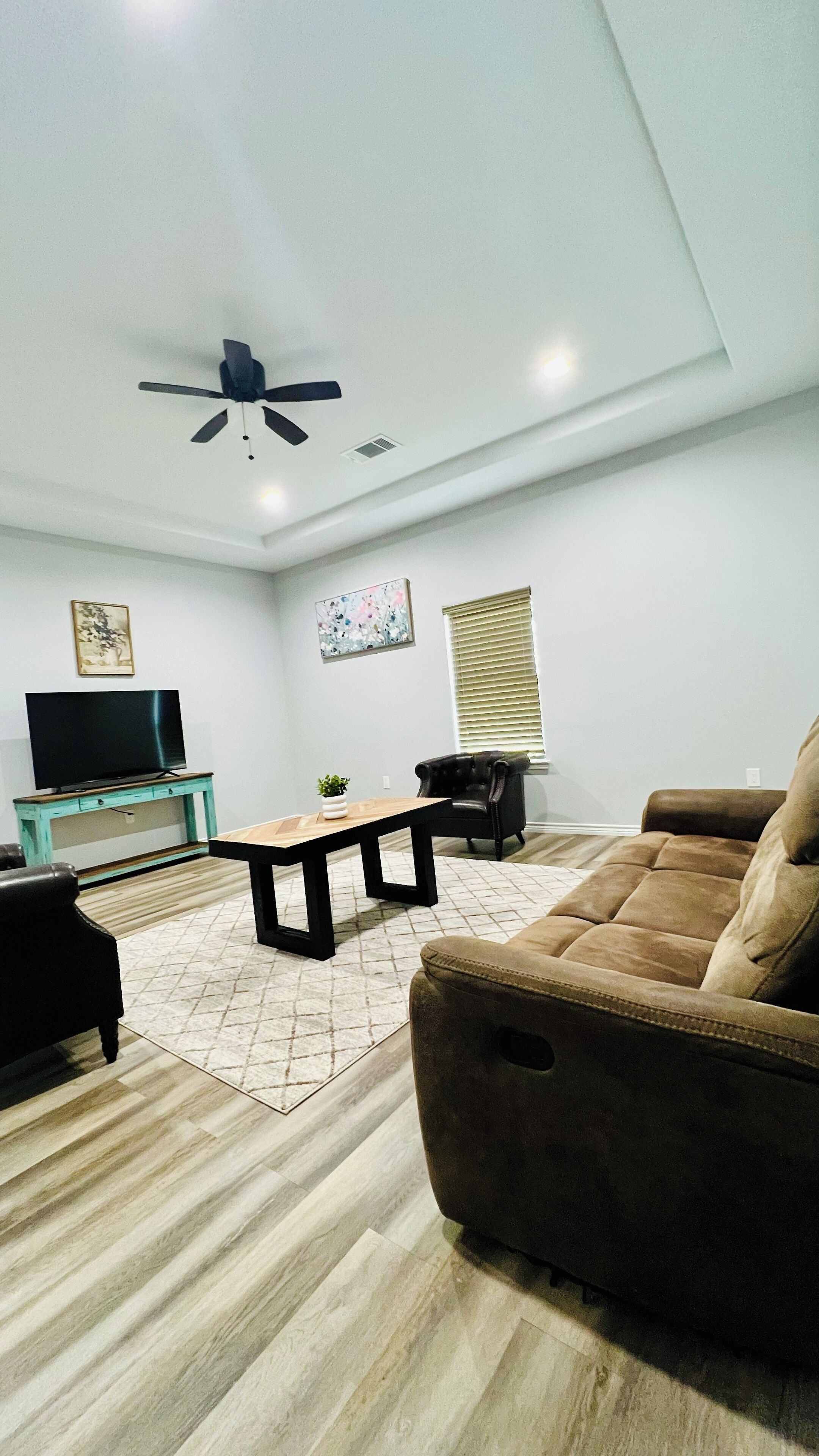 A living room with a sofa, a coffee table, a TV on a stand, a ceiling fan, and a window with blinds.