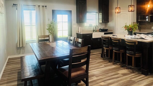 A spacious dining area with a wooden table and chairs, a kitchen with dark cabinets and pendant lighting, and a large window with blinds.