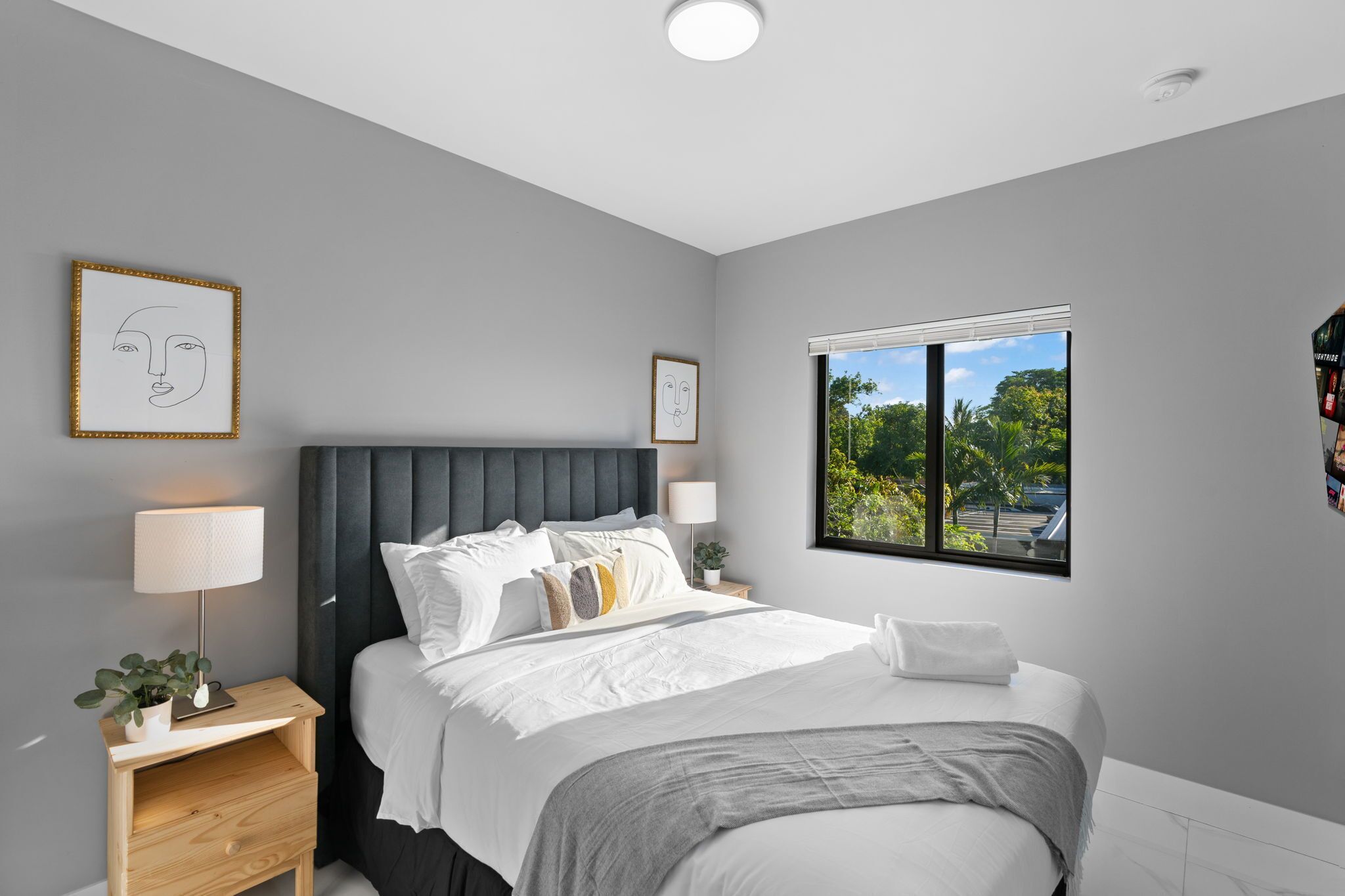 A modern bedroom with a grey upholstered headboard, white bedding, a bedside table with a lamp, and a window with a view of greenery.