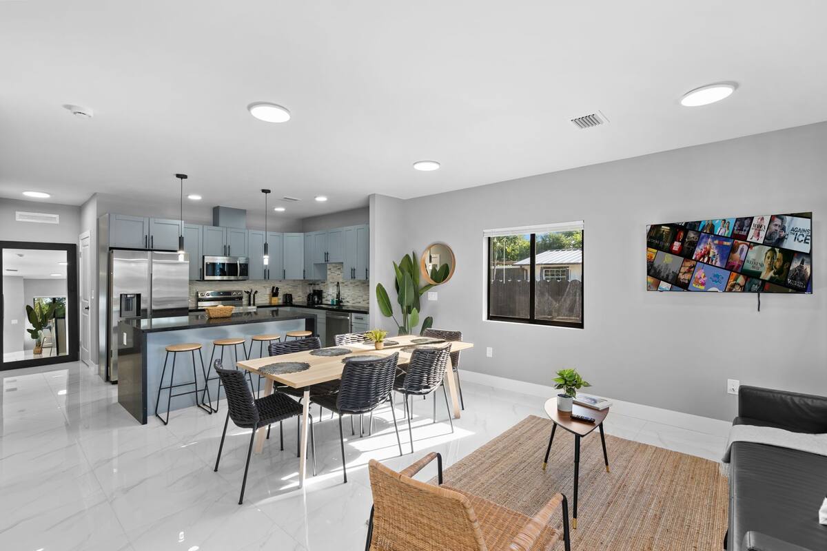 Une cuisine moderne dotée d’appareils électroménagers en acier inoxydable, un coin repas avec une table en bois et des chaises noires, et un grand téléviseur à écran plat fixé au mur.