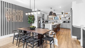 A modern kitchen with a dining area, featuring a wooden table and chairs, a neon sign, and a hanging plant. - Skyline Soundhouse by Avantstay Rooftop Deck & Game Room Near East Nashville & Broadway (Nashville)
