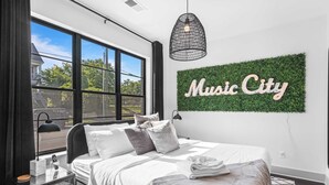 A modern bedroom with a large window, a bed with white bedding, a 'Music City' sign on the wall, and a black pendant light. - Skyline Soundhouse by Avantstay Rooftop Deck & Game Room Near East Nashville & Broadway (Nashville)