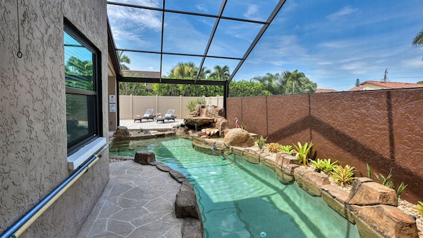 Une piscine bordée de pierres, agrémentée d’une petite cascade, et entourée d’une clôture et de palmiers.