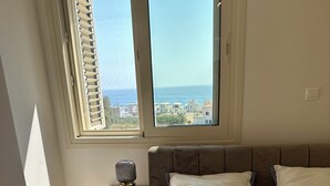 A bedroom with a bed, a nightstand, a lamp, and a view of the sea through large windows.