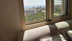 A bedroom with a view of a coastal area, a bed with pillows, a bedside table with a lamp, and a power strip.
